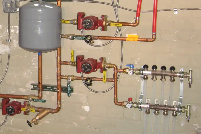 A gray expansion tank and three red circulator pumps on copper pipes leading to a radiant manifold with clear tubing on a basement wall